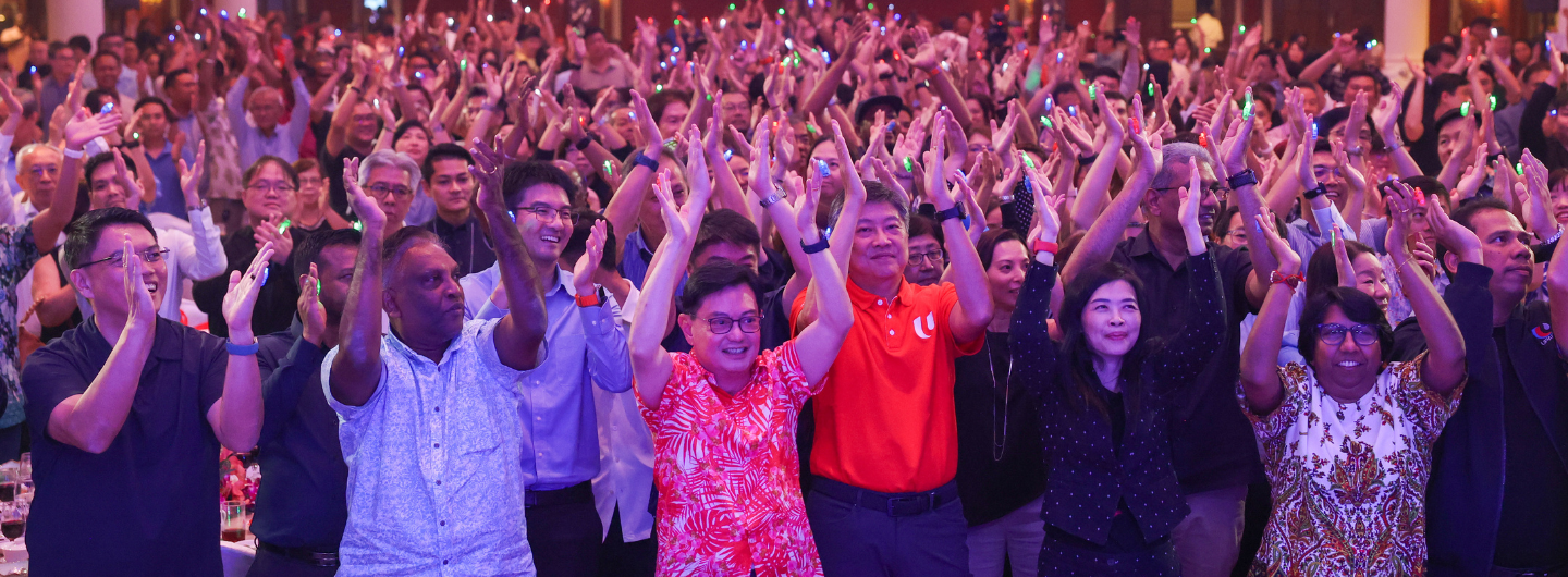 Large crowd clapping with raised hands, some wearing glowing rings, spotlight shines on the event.
